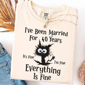 May include: An ivory crew neck t-shirt featuring a black graphic of a dishevelled cat with large blue eyes. The text reads: I've Been Married For 40 Years, It's Fine, I'm Fine, Everything Is Fine. A gold chain necklace rests on the collar area.