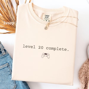 May include: An ivory-coloured t-shirt featuring the text "level 20 complete" and a small game controller graphic. The shirt has a crew neck and a tag that reads "CONCENT CALUKY". A gold necklace is draped across the shirt.