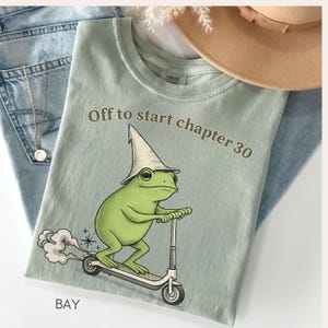 May include: A light green t-shirt with the text "Off to start chapter 30" and a graphic of a green frog wearing a wizard hat and riding a scooter. The shirt is folded on top of blue jeans, with a gold necklace and a tan hat in the background.