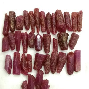 Puede incluir: Una colección de piedras preciosas de rubí en bruto, sin cortar, de diversas formas y tamaños. Las piedras preciosas muestran un tono rojo intenso, con algunas piezas que exhiben una estructura cristalina. La textura rugosa y las formas naturales de los rubíes son visibles.