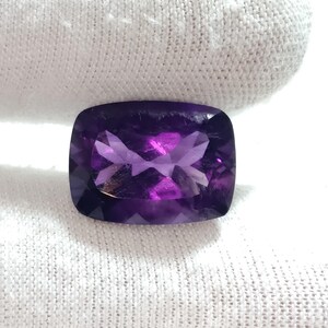 Puede incluir: Una gema de amatista rectangular facetada, de un profundo color p&uacute;rpura. La gema se presenta sobre un fondo de tela blanca, resaltando sus superficies pulidas y los juegos de luz internos. El corte revela un patr&oacute;n complejo.