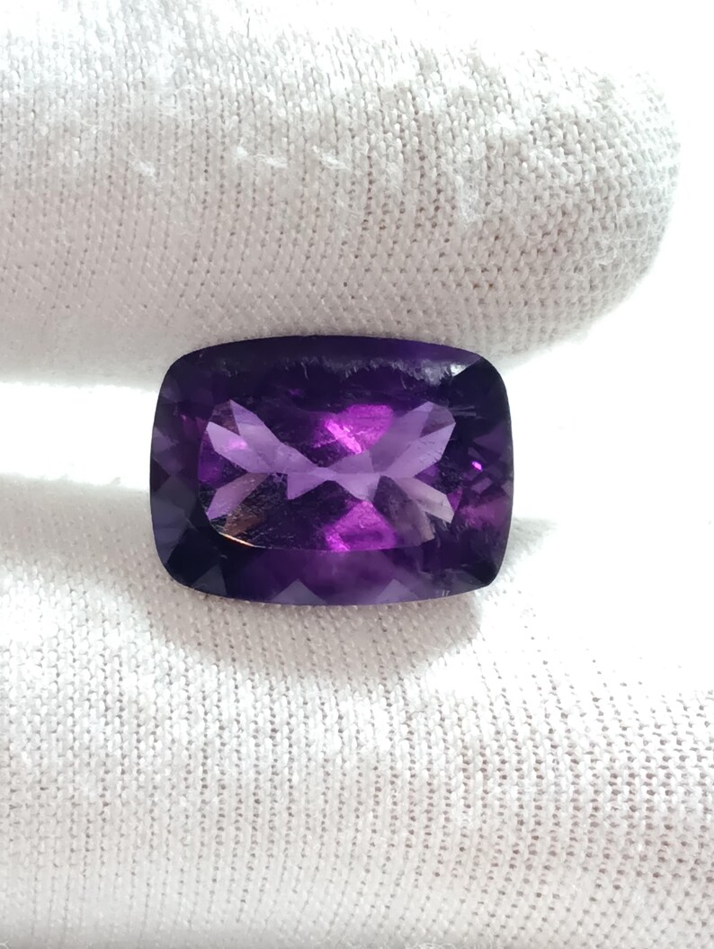Puede incluir: Una gema de amatista rectangular facetada, de un profundo color p&uacute;rpura. La gema se presenta sobre un fondo de tela blanca, resaltando sus superficies pulidas y los juegos de luz internos. El corte revela un patr&oacute;n complejo.