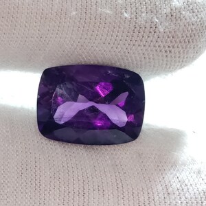 Puede incluir: Una gema de amatista facetada, con corte coj&iacute;n. La gema es de color p&uacute;rpura intenso con reflejos internos de p&uacute;rpura y rosa m&aacute;s claros. La piedra se sostiene sobre un fondo de tela blanca.