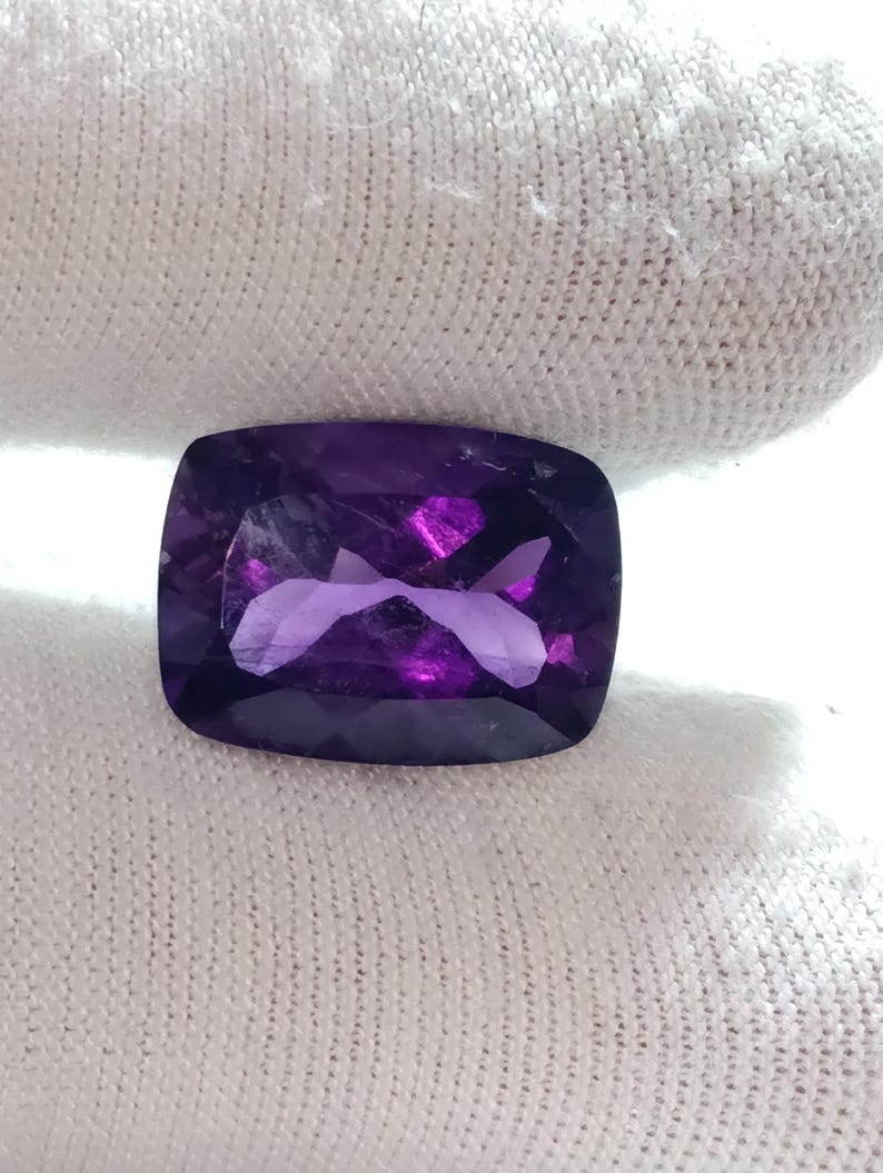 Puede incluir: Una gema de amatista facetada, con corte coj&iacute;n. La gema es de color p&uacute;rpura intenso con reflejos internos de p&uacute;rpura y rosa m&aacute;s claros. La piedra se sostiene sobre un fondo de tela blanca.