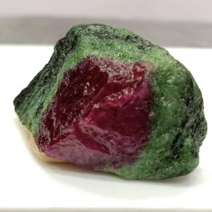 May include: A rough-cut gemstone featuring a vibrant red ruby center surrounded by a green and black matrix. The natural stone has a textured surface and irregular shape, showcasing its raw, unpolished beauty. The gemstone is set against a white background.
