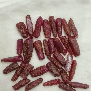 Puede incluir: Una colección de piedras preciosas de rubí en bruto de varias formas y tamaños. Las piedras en bruto muestran un tono rojo intenso, algunas exhiben un tono ligeramente más claro, violáceo. Las piedras preciosas están dispersas sobre una superficie blanca.