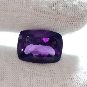 Puede incluir: Una piedra preciosa de amatista tallada en coj&iacute;n. La gema es de color p&uacute;rpura intenso con reflejos internos. La piedra tiene esquinas redondeadas y forma rectangular. La piedra preciosa est&aacute; sobre un fondo blanco.