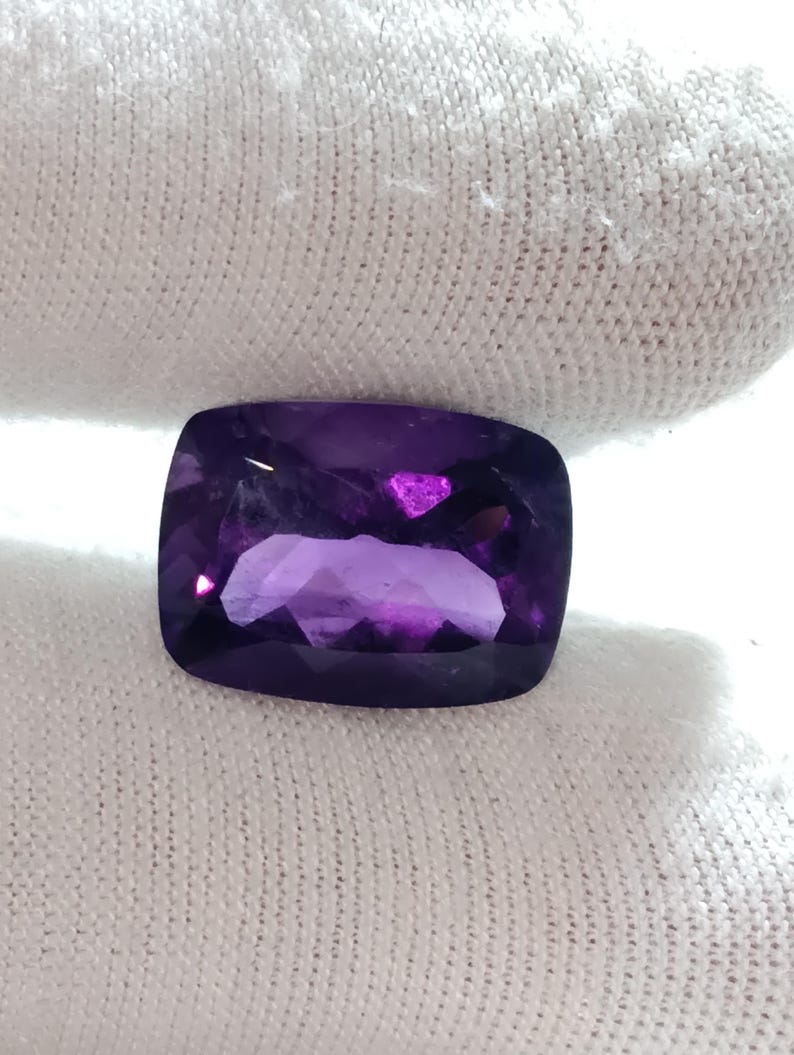 Puede incluir: Una piedra preciosa de amatista tallada en coj&iacute;n. La gema es de color p&uacute;rpura intenso con reflejos internos. La piedra tiene esquinas redondeadas y forma rectangular. La piedra preciosa est&aacute; sobre un fondo blanco.