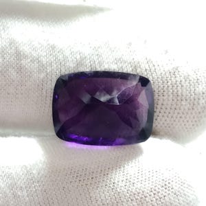 Puede incluir: Una piedra preciosa de amatista tallada en coj&iacute;n. La gema es de color p&uacute;rpura intenso con una superficie pulida. La piedra preciosa se coloca sobre un fondo blanco, resaltando su color y claridad. El corte de la piedra permite que la luz se refleje, creando un efecto brillante.
