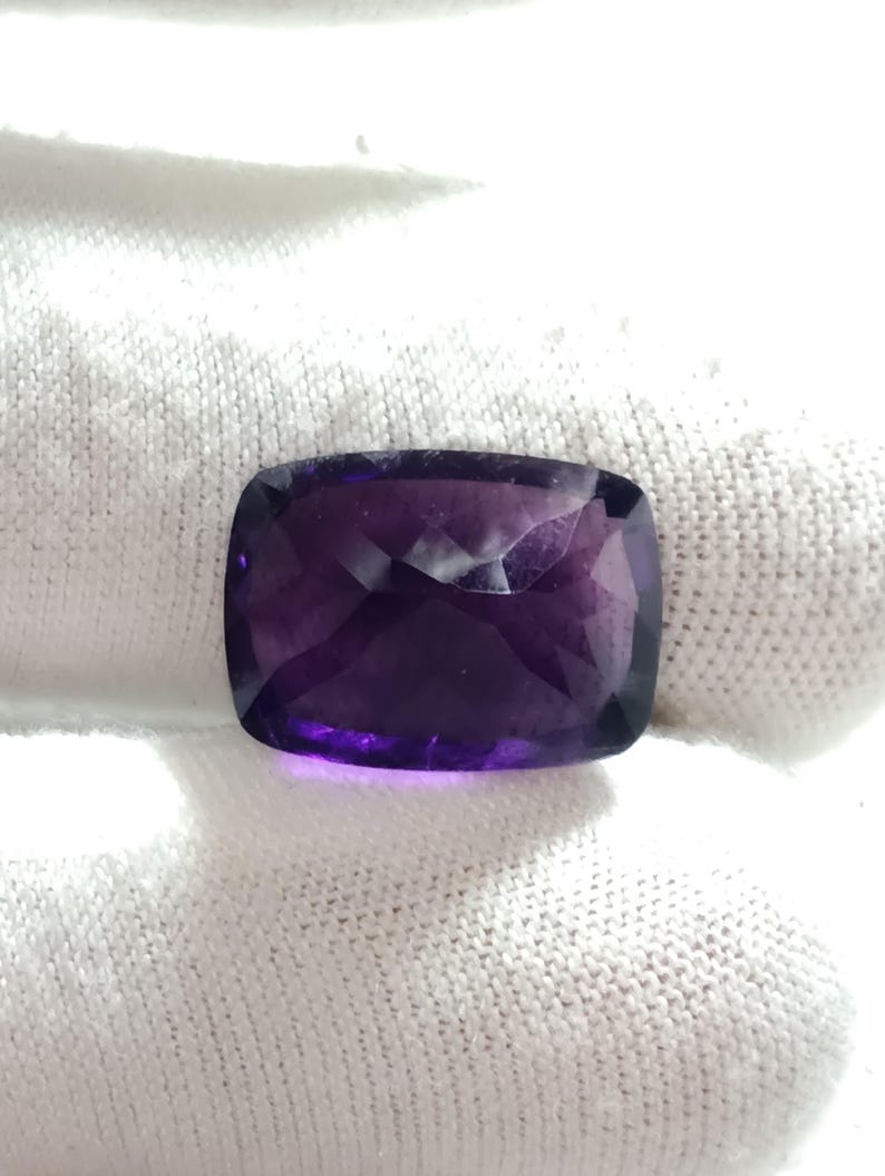 Puede incluir: Una piedra preciosa de amatista tallada en coj&iacute;n. La gema es de color p&uacute;rpura intenso con una superficie pulida. La piedra preciosa se coloca sobre un fondo blanco, resaltando su color y claridad. El corte de la piedra permite que la luz se refleje, creando un efecto brillante.