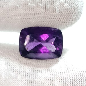 Puede incluir: Una piedra preciosa de amatista con talla coj&iacute;n. La gema es de color p&uacute;rpura intenso con facetas que reflejan la luz. La piedra est&aacute; sobre un fondo blanco texturizado.