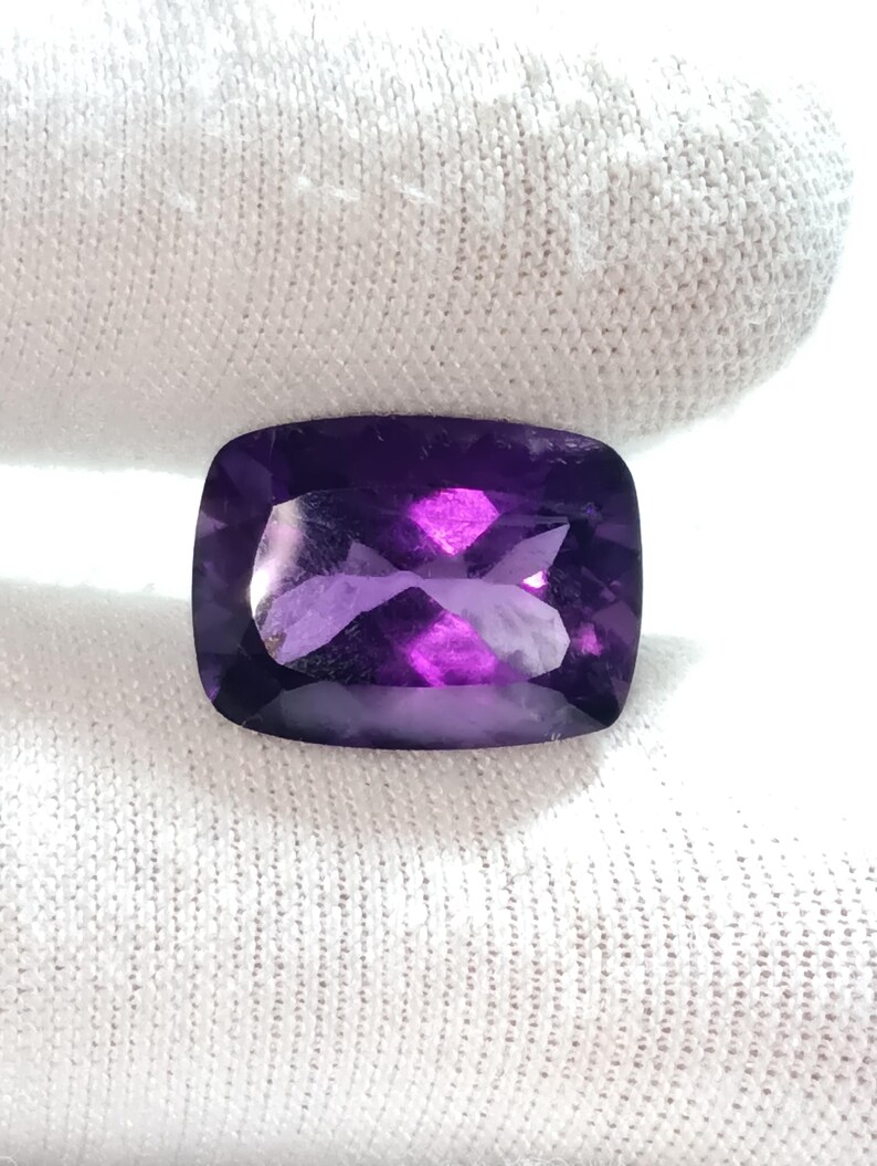 Puede incluir: Una piedra preciosa de amatista con talla coj&iacute;n. La gema es de color p&uacute;rpura intenso con facetas que reflejan la luz. La piedra est&aacute; sobre un fondo blanco texturizado.