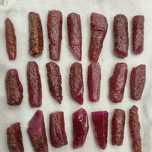 May include: A collection of raw, uncut ruby gemstones. The rubies vary in shape and size, displaying a deep red hue with textured surfaces. The gemstones are arranged on a white cloth, showcasing their natural beauty.