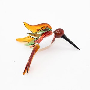 May include: A glass hummingbird figurine with brown, orange, green, and white glass. The hummingbird is in flight with its wings spread out.