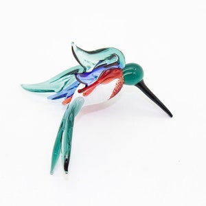 May include: A glass hummingbird figurine with a green, blue, red, and white color scheme. The hummingbird is in flight with its wings spread out.