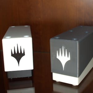 Könnte beinhalten: Zwei rechteckige Kartenaufbewahrungsboxen, eine weiße und eine graue, jeweils mit einem schwarz-weißen Emblem. Die Boxen haben ein zweifarbiges Design und sind wahrscheinlich für Sammelkartenspiele.