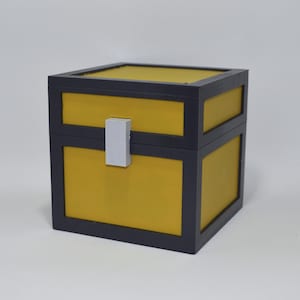 Op de afbeelding: Een vierkante kist met een gele binnenkant en een donkergrijs frame. De kist heeft een witte rechthoekige sluiting in het midden. Het ontwerp doet denken aan een videogamekist.