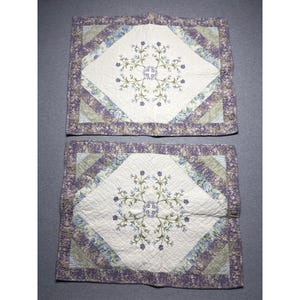 May include: Two rectangular quilted pillow shams featuring a central white diamond panel with embroidered blue and green floral motifs. The borders consist of diagonal strips in shades of purple, blue, and green with a floral pattern.