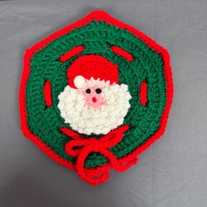Può includere: Un sottopentola natalizio fatto a mano, all'uncinetto, con Babbo Natale. Il sottopentola ha una base verde con bordi e dettagli rossi. La faccia di Babbo Natale è bianca con un naso rosa e occhi neri, con un cappello rosso. Un fiocco rosso è in basso.