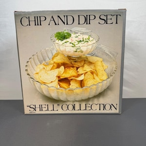 Puede incluir: Un juego de patatas fritas y salsa en una caja. La caja dice "CHIP AND DIP SET" y "SHELL COLLECTION". El juego incluye un cuenco grande y transparente en forma de concha lleno de patatas fritas y un cuenco más pequeño con salsa.