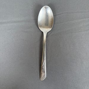 Puede incluir: Una cuchara plateada con un cuenco ovalado y un mango largo y delgado. El mango presenta un diseño de trigo grabado cerca de la base. La cuchara está sobre un fondo gris.