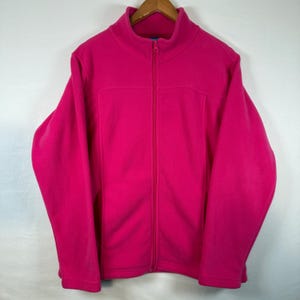 May include: A vibrant fuchsia fleece jacket with a full-length zipper and a high collar. The jacket is hanging on a wooden hanger against a plain white background. The jacket has two front pockets and a soft, textured appearance.