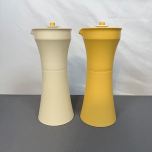 Può includere: Due barattoli da cucina color crema e giallo oro vintage. Ogni barattolo ha un corpo cilindrico con una base svasata e un piccolo beccuccio. I coperchi sono piatti con una piccola maniglia quadrata. I barattoli sono di plastica.