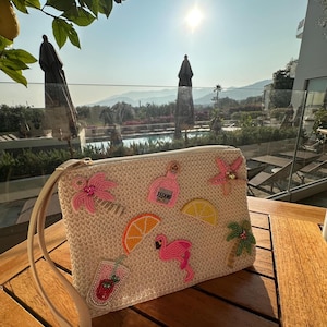 May include: Cream-coloured clutch bag with a woven texture, decorated with beaded tropical embellishments. Features a pink flamingo, orange slice, and a cocktail. Includes a wrist strap.
