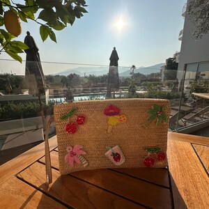 May include: A rectangular straw clutch bag with beaded decorations of cherries, a cocktail, a palm tree, a flower, and a strawberry drink. The bag has a wrist strap and is on a wooden table.