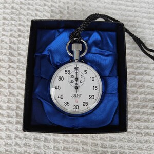 May include: A silver stopwatch with a black cord, presented in a black box with blue satin lining. The stopwatch face is white with black numerals and hands, and the brand name "DOLMY" is visible.