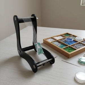 May include: A black wooden bead loom with a partially woven beaded project. A wooden tray with various coloured beads and small containers of beads are also on the table. A needle is also present.
