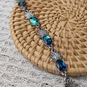 Puede incluir: Pulsera plateada con dijes de piña con centros de concha de abulón iridiscente. La pulsera se muestra sobre una superficie tejida de color marrón claro, mostrando el tema tropical y el diseño de la joya.