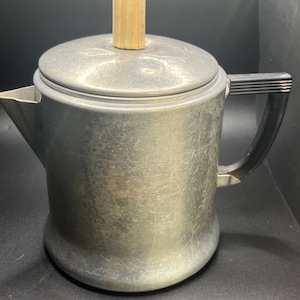 Könnte beinhalten: Eine silberfarbene Vintage-Kaffeekanne mit Deckel und Holzgriff. Die Kanne hat einen schwarzen Griff und einen Ausguss. Das Metall hat ein abgenutztes Aussehen, was auf Alter und Gebrauch hindeutet. Ein klassischer Küchenartikel.