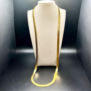 May include: A gold-colored herringbone chain necklace displayed on a mannequin. The necklace is flat and flexible, with a smooth, reflective surface. The chain is draped in a long, elegant style.