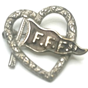 May include: A vintage silver-toned pin in the shape of a heart with an ornate, swirling design. A banner with the letters "F.F.F." is featured in the center, along with a small flag. The pin has a classic, antique aesthetic.