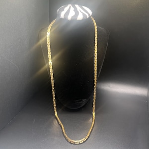 May include: A gold-colored necklace with a rectangular link design. The necklace is displayed on a black jewelry bust with a zebra-striped top. The background is black.
