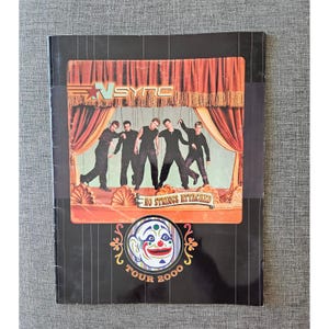 May include: A black and red *NSYNC "No Strings Attached" Tour 2000* album cover. The cover features a stage design with the band members in black outfits. The bottom of the cover has a clown face design.