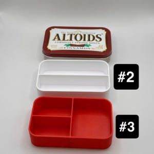 May include: An Altoids tin with a red and gold label sits above two plastic organizers. The top organizer is white with two compartments, and the bottom organizer is red with three compartments. The text on the tin reads "Altoids Cinnamon".