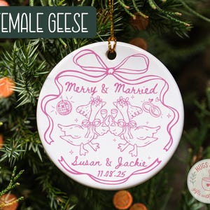 May include: White ceramic ornament with a pink illustration of two geese toasting, with the text "Merry & Married" and names "Susan & Jackie" with a date. The ornament is hanging on a Christmas tree.