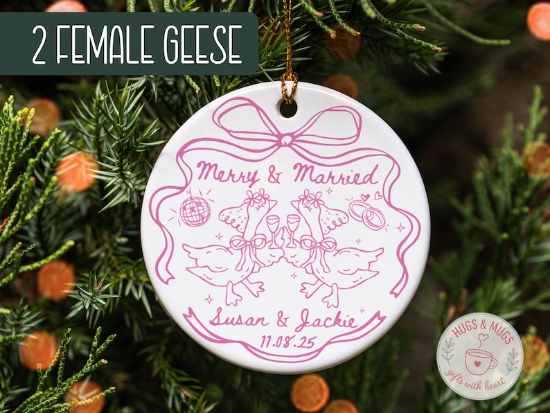 May include: White ceramic ornament with a pink illustration of two geese toasting, with the text "Merry & Married" and names "Susan & Jackie" with a date. The ornament is hanging on a Christmas tree.