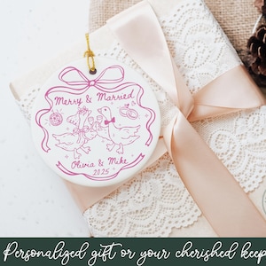 May include: A white ceramic ornament with a pink design featuring two geese toasting with champagne glasses, a disco ball, and wedding rings. The text reads "Merry & Married", "Olivia & Mike", and "2025". The ornament hangs on a gold string.