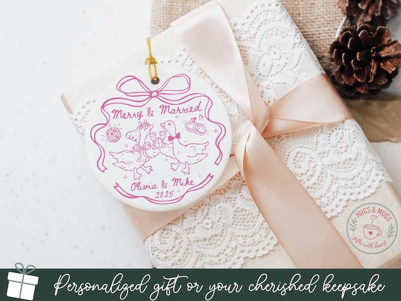 May include: A white ceramic ornament with a pink design featuring two geese toasting with champagne glasses, a disco ball, and wedding rings. The text reads "Merry & Married", "Olivia & Mike", and "2025". The ornament hangs on a gold string.