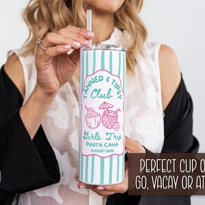 May include: A white and teal striped tumbler with a lid and straw. The tumbler has a pink label that reads "Tanned & Tipsy Club Girls Trip Punta Cana August 2026". The tumbler is held by a person.