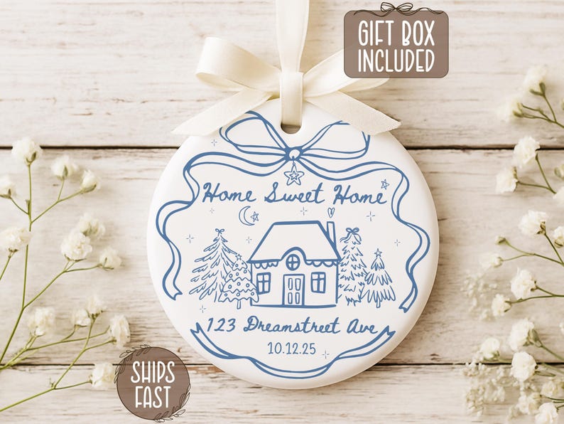 May include: White ceramic ornament with blue illustrations and text. The ornament features a house, trees, and the words "Home Sweet Home" and "123 Dreamstreet Ave". Includes a cream-colored ribbon for hanging. The words "Gift Box Included" are also visible.
