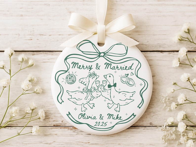 May include: White ceramic wedding ornament with a green illustration of two geese toasting with champagne glasses. The ornament reads "Merry & Married" with the names "Olivia & Mike" and the year "2025". A white ribbon is attached.