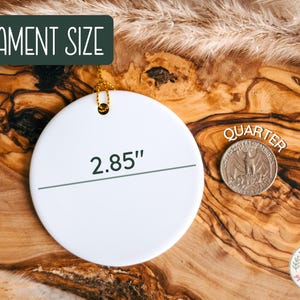 May include: A white, round ornament with a gold string for hanging. The ornament is 2.85 inches in diameter. The text "Ornament Size" is in a green box. A quarter coin is next to the ornament.