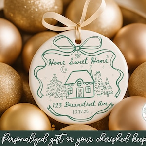 May include: A round, white ceramic ornament with a green illustration of a house, trees, and the text "Home Sweet Home." The ornament is surrounded by gold Christmas ornaments and has a cream-colored ribbon. The text "123 Dreamstreet Ave" and "10.12.25" are also included.