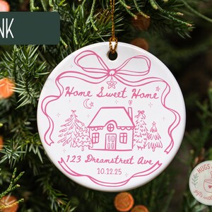 May include: A white ceramic ornament with a pink illustration of a house, trees, and the text "Home Sweet Home" and "123 Dreamstreet Ave 10.12.25". The ornament is hanging on a Christmas tree.