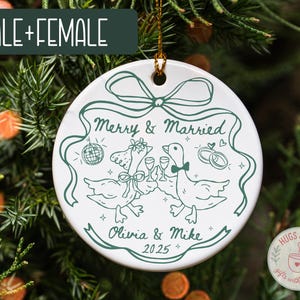 May include: White ceramic Christmas ornament with a green illustration of two geese toasting with champagne glasses. The ornament reads "Merry & Married" with the names "Olivia & Mike" and the year "2025".