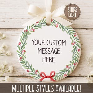 May include: A white, round ornament with a cream-colored ribbon. The ornament features a green and red wreath design around the edge and the text "YOUR CUSTOM MESSAGE HERE". A red bow is at the bottom. The text "MULTIPLE STYLES AVAILABLE!" is at the bottom.